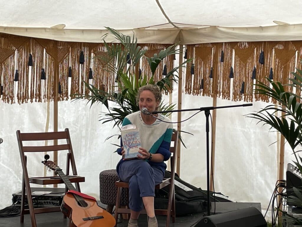 Catrina Davies reading from the start of her third book based on the recollections of Hedley Ralph Collard. Why Stories About Climate Change need a hook. She sits in a small marquee on a seat, with a guitar to the right of her.